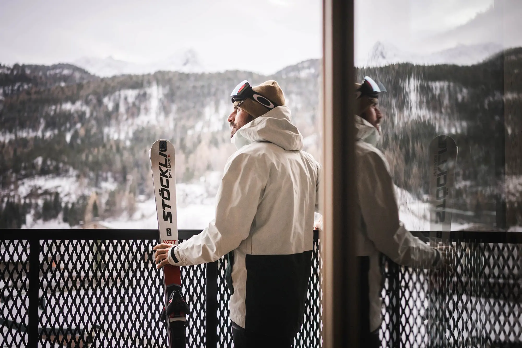 Eine Person in Skiausrüstung steht mit Skiern in der Hand auf einem Balkon und blickt auf schneebedeckte Berge, deren Spiegelbild sich im Glas spiegelt.