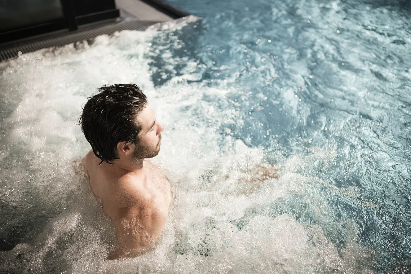 Mann entspannt sich in einem sprudelnden, teilweise untergetauchten Whirlpool, während sich helles Sonnenlicht im Wasser spiegelt.