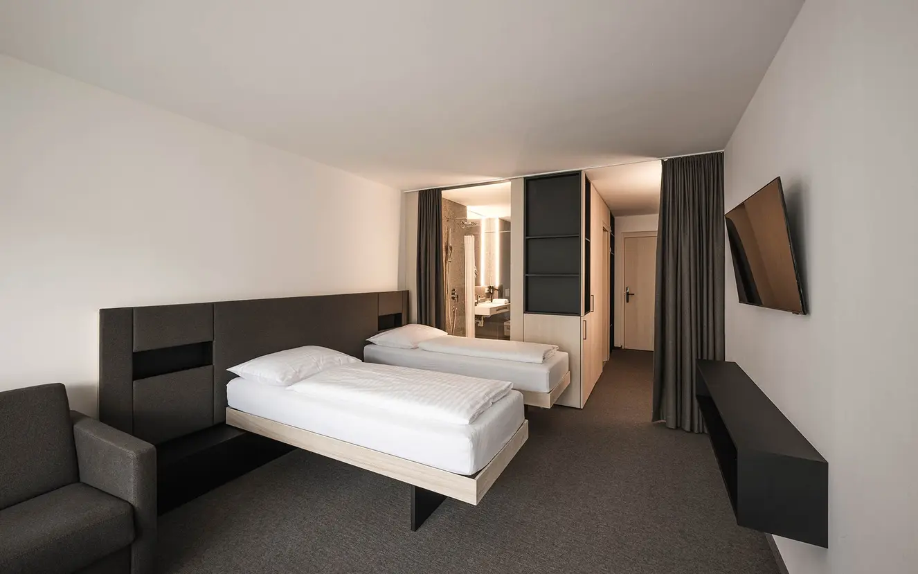 Minimalistisches Hotelzimmer mit zwei Einzelbetten, einem an der Wand montierten Fernseher, einem grauen Sofa und einem modernen offenen Badezimmer.
